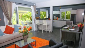Un salon moderne avec un canapé, une table basse et des fauteuils. On y trouve également un coin repas avec une table dressée pour deux personnes. La cuisine est visible, avec ses placards et son réfrigérateur.