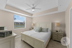 Un dormitorio con cama, mesitas de noche, ventilador de techo, ventana con persianas y una pequeña mesa con lámpara.