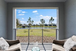Un balcón con muebles de mimbre, una mesa de vidrio y vistas a un lago con casas al fondo.
