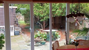 Une terrasse couverte avec une table et des chaises, un luminaire suspendu et une vue sur un jardin avec des plantes et une terrasse en bois.