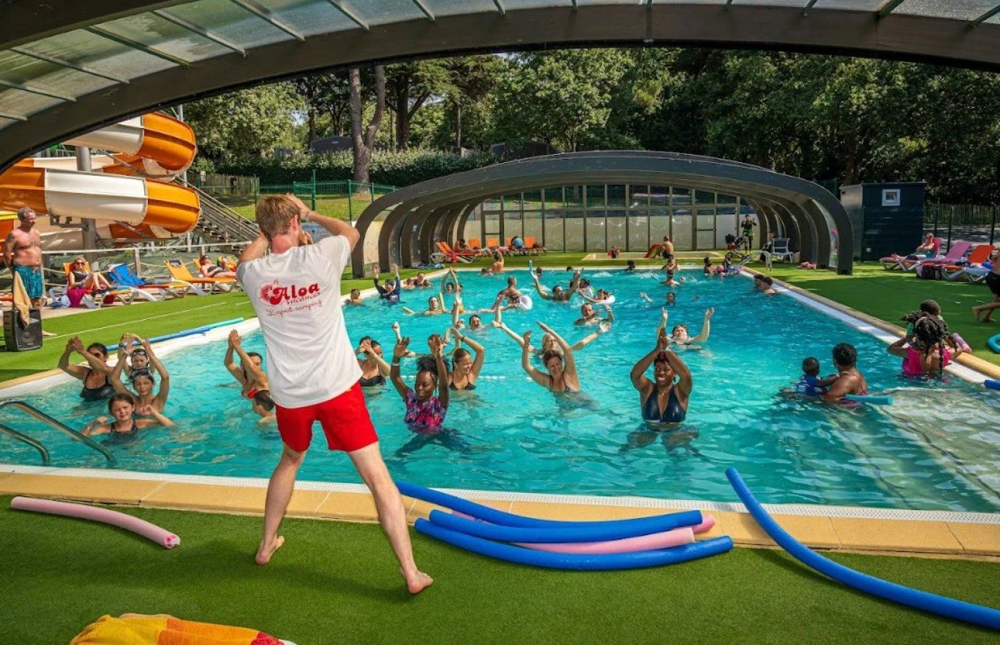 An outdoor swimming pool with a covered area, surrounded by people engaging in water activities.