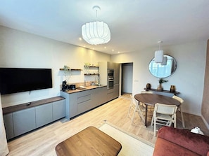 A modern kitchen with a flat-screen TV, built-in oven, and a dining area with a round table and chairs. - L'appart Rémois | Balcon | Centre-ville (Reims)