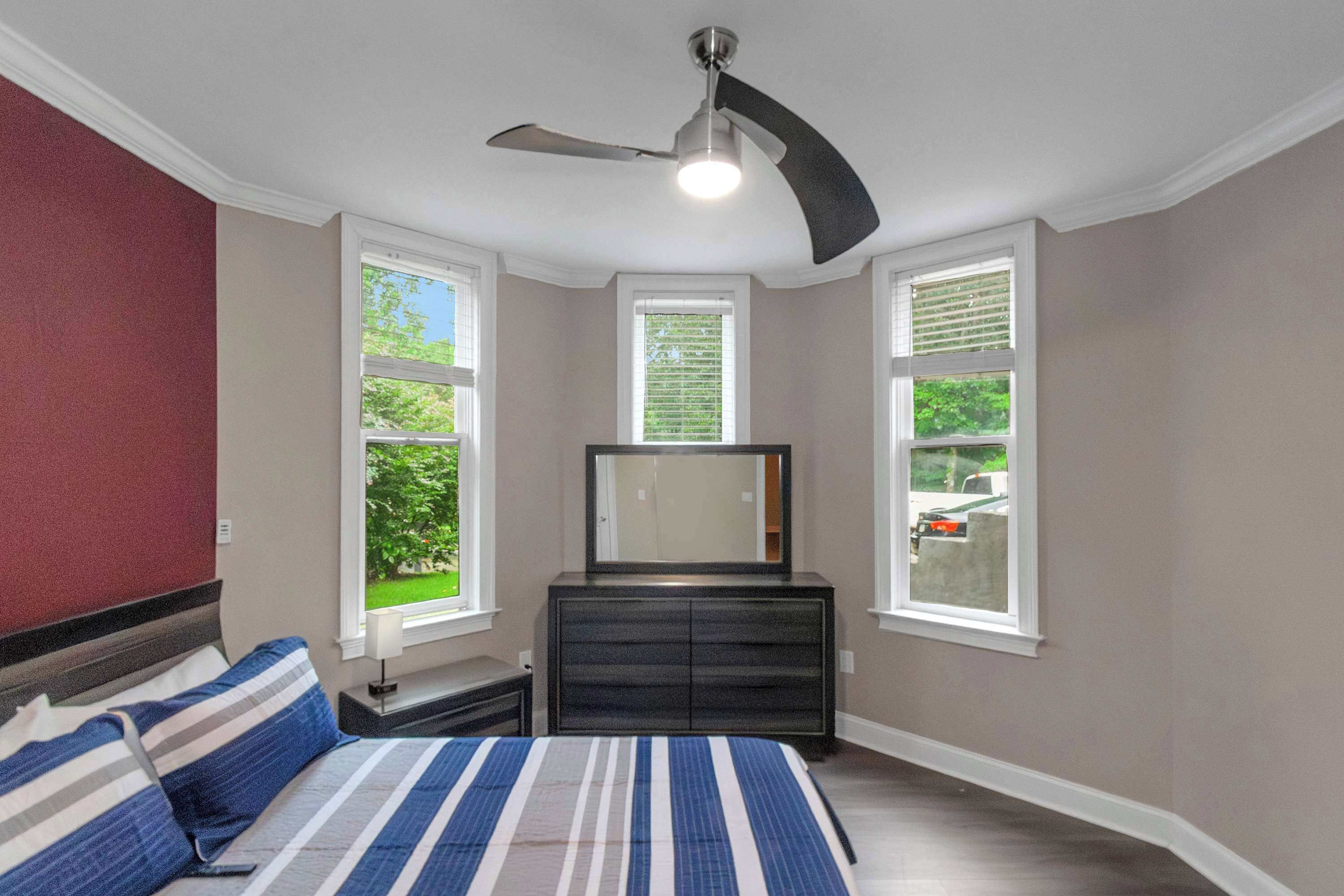 A bedroom with a bed, a dresser, a ceiling fan, and windows with views of greenery.