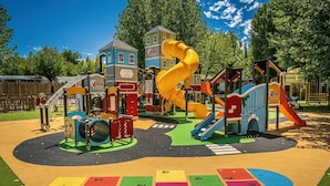 Ein Spielplatz mit Rutschen und Klettergerüsten, umgeben von einem Zaun und Bäumen.