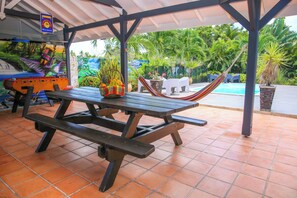 A covered patio area with a picnic table, a hammock, and a pool in the background. - Les NUITS GALANTES - Apartment LANTANA (Saint-François)