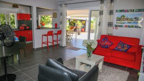 A modern living room with a red sofa, black leather chairs, and a white coffee table. There is a kitchen area with red cabinets and a fan.