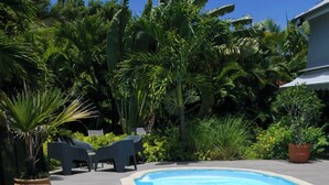 A poolside area with white lounge chairs, a small round table with drinks, and lush greenery in the background.