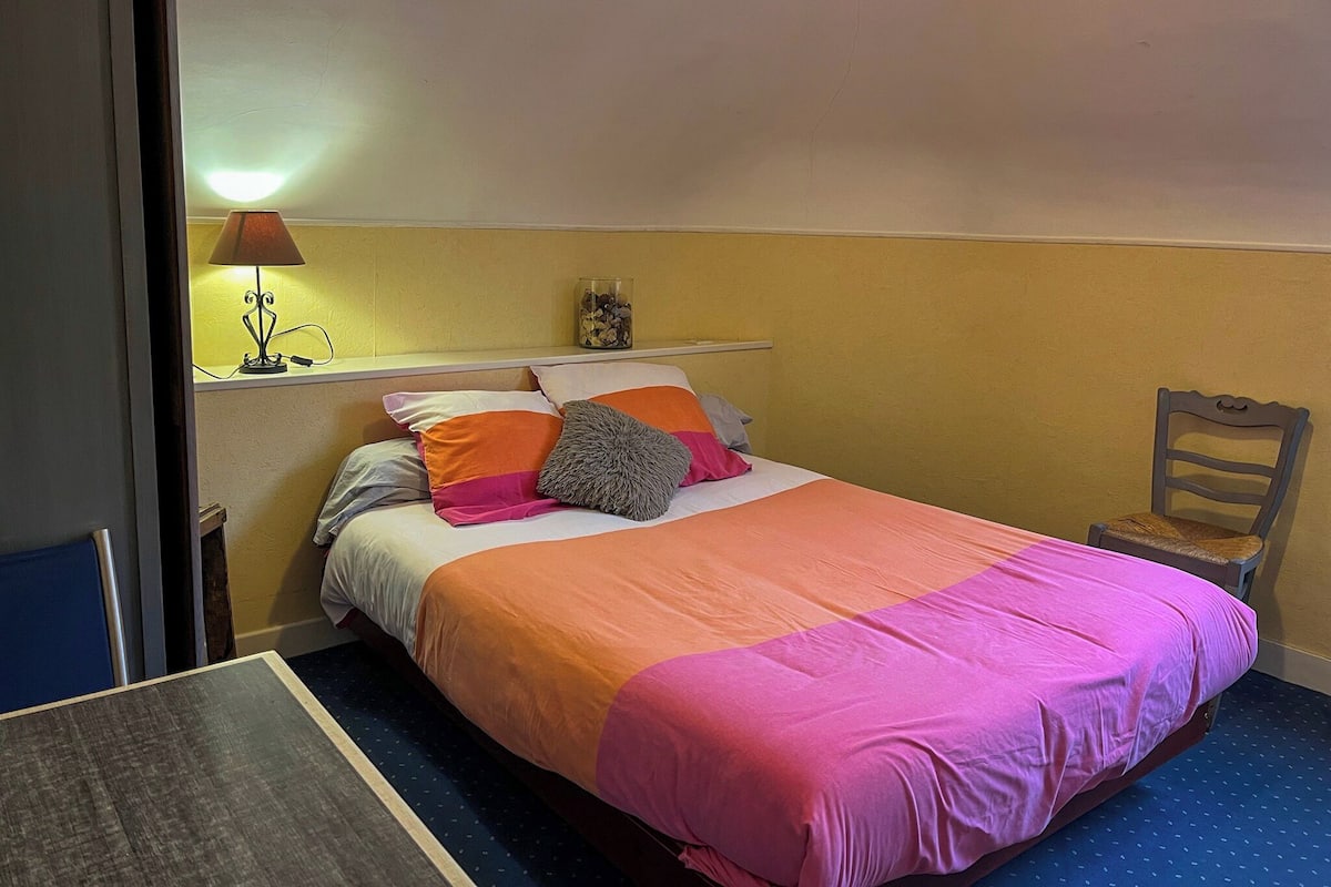 A bedroom with a bed, a nightstand, a lamp, a chair, and a shelf with decorative items.