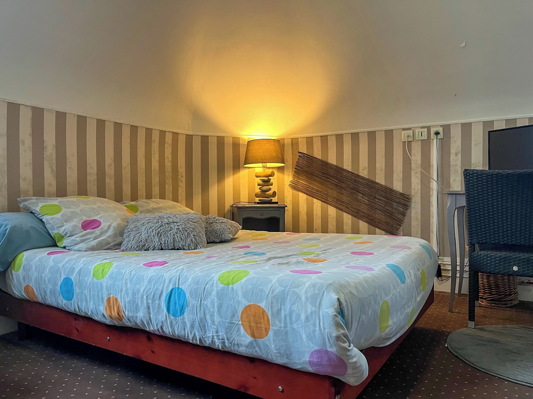 A bedroom with a bed, pillows, a lamp, a chair, and striped wallpaper.