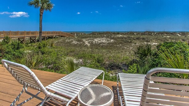 A wooden deck with metal chairs and a round table overlooking a beach with a wooden boardwalk and a palm tree. - 3rd Floor Villa w/ Ocean Views & Amenities (Hilton Head Island)