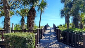 A wooden boardwalk with metal railings, surrounded by lush greenery and palm trees, leading towards a clear blue sky. - 3rd Floor Villa w/ Ocean Views & Amenities (Hilton Head Island)