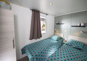A bedroom with a bed, a window with curtains, a bedside table, and a wall-mounted shelf with containers. - ZEcamping parc aquatique animations mobil-home TV LV 35m2 6 pers (Saint-Michel-Chef-Chef)