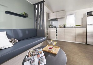 Een moderne woonkamer met een grijze bank, een salontafel met tijdschriften, en een keukenhoek met licht houten kasten en een roestvrijstalen koelkast.
