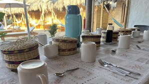 Una mesa dispuesta con tazas blancas, un termo azul y cestas tejidas, con un techo de paja y sillones de descanso al fondo.
