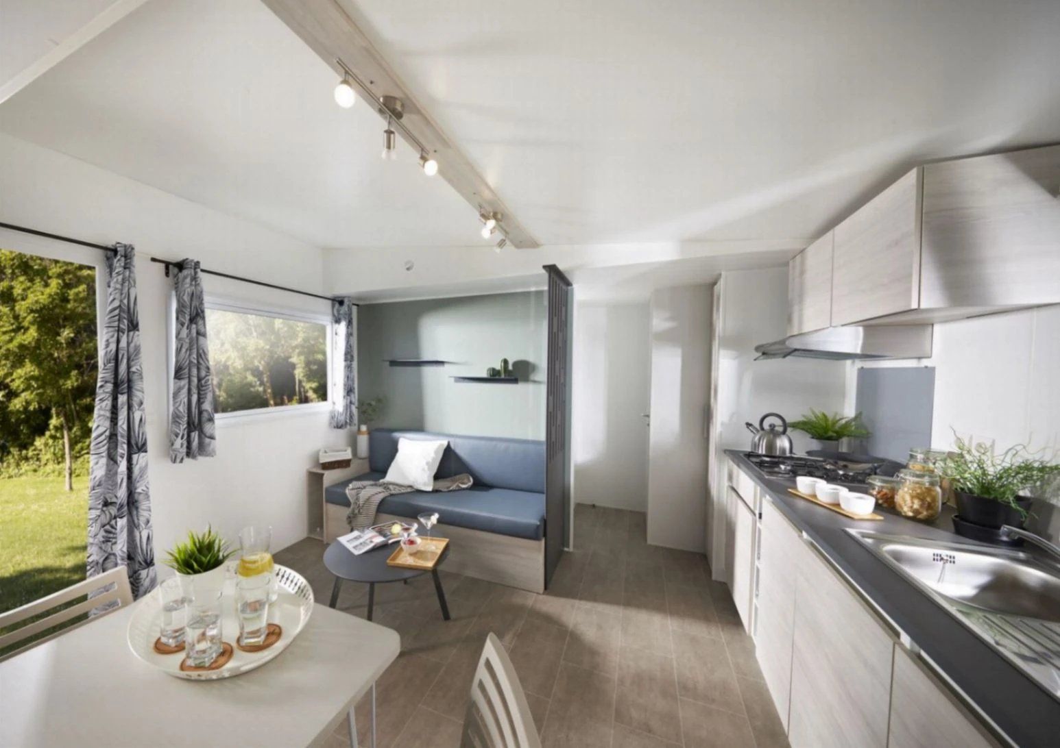 Een ruimtelijk ingerichte leefruimte met een kitchenette, een eethoek met een tafel en stoelen, een zithoek met een bank, en een raam met gordijnen.