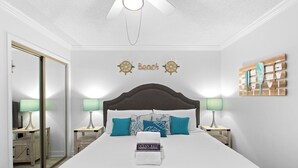 A bedroom with a large bed, two bedside tables with lamps, a mirror, a ceiling fan, and a wall with a nautical-themed decoration.