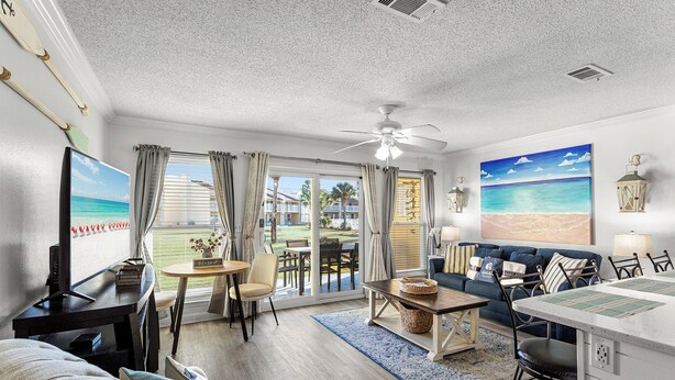 A spacious living area with a large TV, a dining table, and a sofa. There is a ceiling fan and a view of the outdoors through the windows.