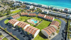 An aerial view of a resort with a central pool, surrounded by buildings with red-tiled roofs, and a beach in the background.