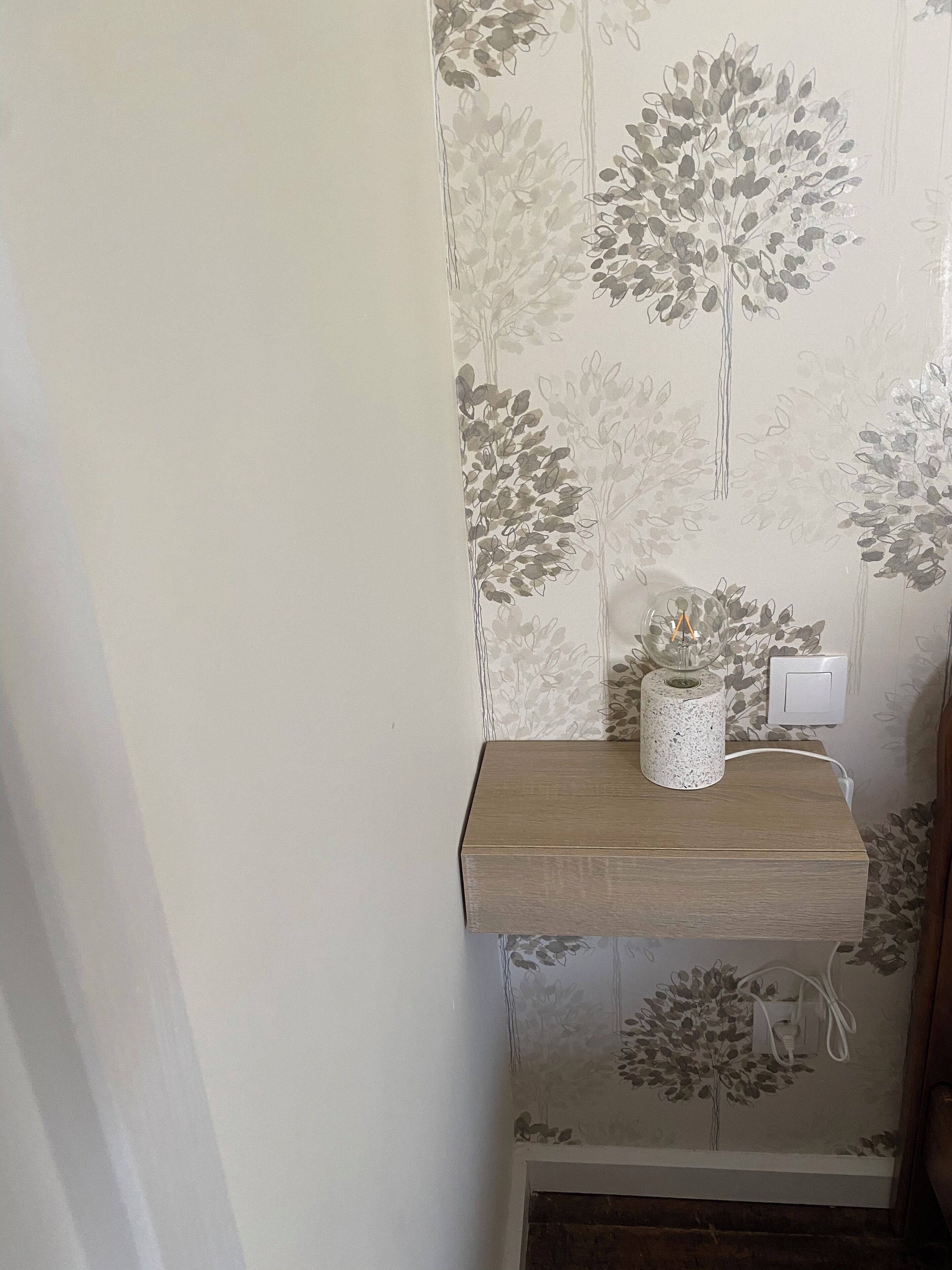 A bathroom with floral wallpaper, a wooden shelf with a vase and a roll of toilet paper, and a power outlet.
