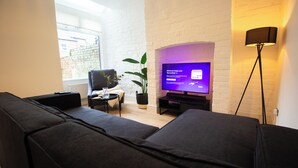 A modern living room with a flat-screen TV, a black sofa, a small side table, a potted plant, and a floor lamp. - Comfy 4 Bed, Quiet Stay, Near the City Center, Free Parking & Speedy WIFI  (Manchester)