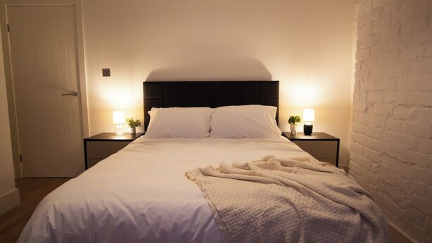 A hotel room with a large bed, two bedside tables with lamps, a white brick wall, and a door.