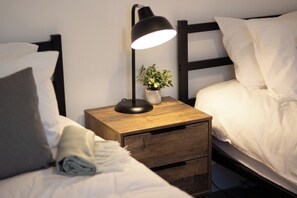 Une chambre à coucher avec une table de chevet en bois, une lampe, une petite plante et un lit recouvert de linge de lit blanc.