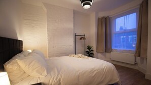 A bedroom with a bed, a bedside table, a radiator, a window with curtains, and a wall-mounted plant.