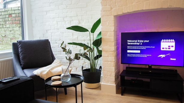 A living room with a black leather sofa, a small round table with a plant and a vase, and a flat-screen TV displaying a welcome message. - Comfy 4 Bed, Quiet Stay, Near the City Center, Free Parking & Speedy WIFI  (Manchester)