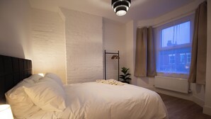 A bedroom with a bed, a nightstand, a wall-mounted radiator, and a window with curtains. - Comfy 4 Bed, Quiet Stay, Near the City Center, Free Parking & Speedy WIFI  (Manchester)