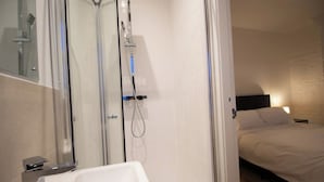 A modern bathroom with a glass shower enclosure, a white bathtub, and a bed with white bedding in the adjacent room.