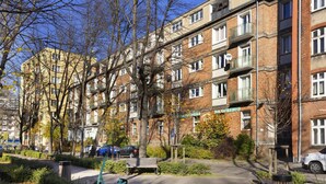 Un immeuble résidentiel à plusieurs étages, doté de balcons, d’une allée pavée et d’un petit espace vert aménagé avec des bancs et des arbres.