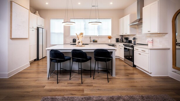 Una cocina moderna con una isla central, taburetes negros, muebles blancos y un amplio ventanal que proporciona luz natural.
