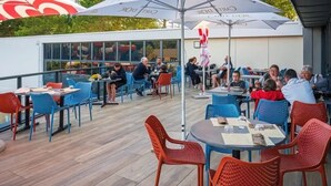An outdoor dining area with tables and chairs, a flag, and a building with windows in the background. - ZEcamping parc aquatique chiens acceptés club enfant locatif TV LV (Saint-Hilaire-de-Riez)