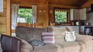 Interior acogedor de una cabaña con techo de madera, un sofá con cojines, una zona de comedor con mesa y sillas, y una cocina con electrodomésticos básicos.