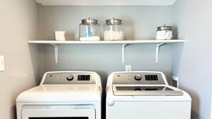 A laundry room with two front-loading washing machines and a shelf above containing jars and a cup. - Your Little Guest House (Kennewick)