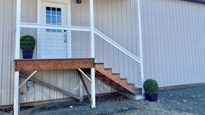 A building with a wooden staircase leading to a white door, a small porch with a railing, and a potted plant on the ground. - Your Little Guest House (Kennewick)