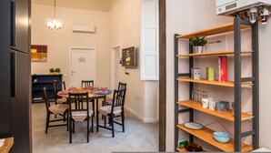 Un coin repas avec une table ronde, des chaises et un lustre. Il y a également un coin cuisine équipé d’un réfrigérateur, d’une étagère contenant divers objets et d’un téléviseur fixé au mur.