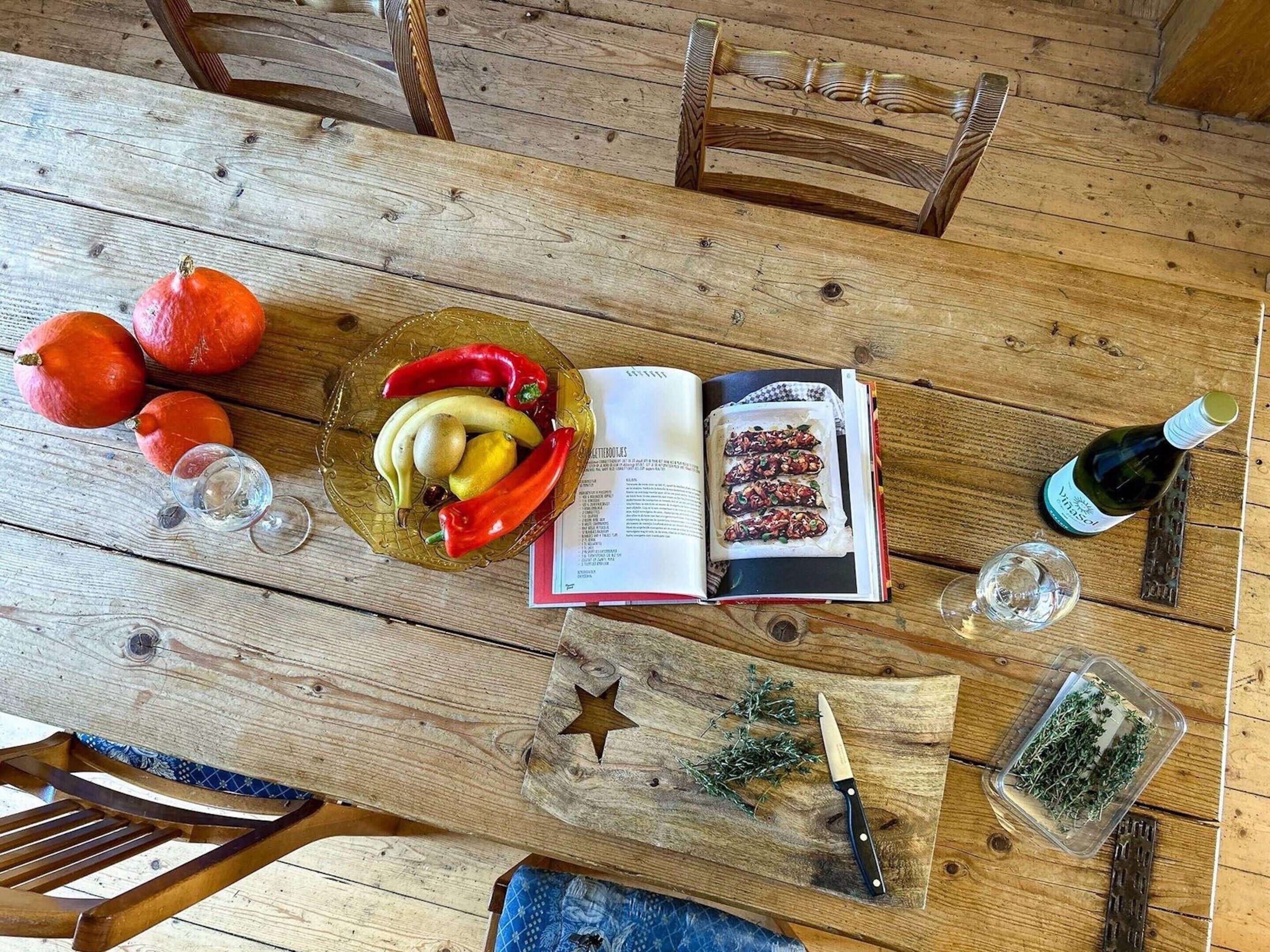 A wooden dining table with a book, a glass of water, a bottle of wine, and a bowl of fruit.