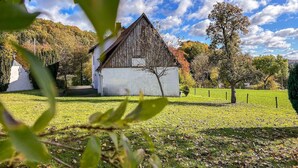 A rustic barn with a white exterior and a wooden roof, surrounded by a grassy field and trees, under a partly cloudy sky.
