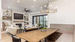 A modern living room with a wooden dining table, white sofas, a fireplace, and large windows.