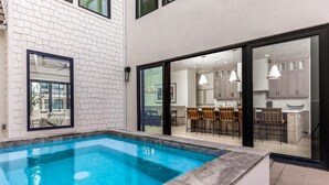 A modern house with a swimming pool, a patio with outdoor furniture, and a kitchen visible through glass doors.
