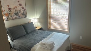 Een slaapkamer met een bed, kussens, een bedlampje, een raam met zonwering en een schilderij aan de muur.