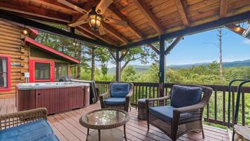 A covered deck with wicker furniture, a hot tub, and a view of the surrounding forest.