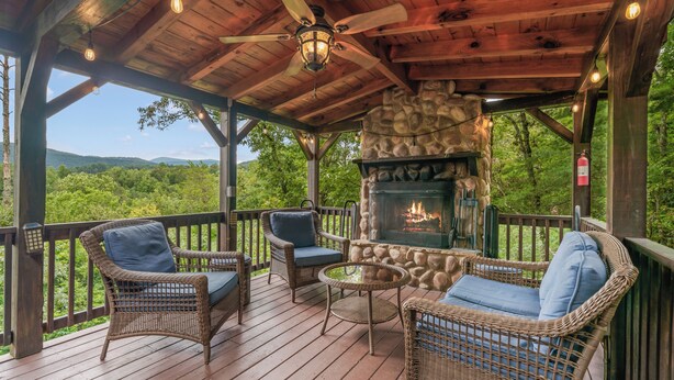 A covered outdoor seating area with wicker furniture, a stone fireplace, and a view of the surrounding forest. - Views for Days-Big Game Room-Hot Tub-3 King Beds! (Blue Ridge)