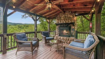A covered outdoor seating area with wicker furniture, a stone fireplace, and a view of the surrounding forest.