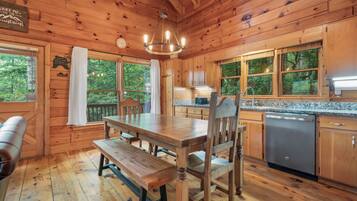 A wooden cabin interior with a dining table, benches, a chandelier, wooden cabinets, and a kitchen area.