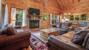 A cozy cabin interior with wooden walls, leather sofas, a stone fireplace, and a kitchen area. - Views for Days-Big Game Room-Hot Tub-3 King Beds! (Blue Ridge)