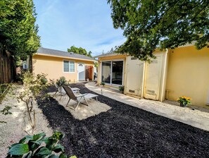 Un patio trasero residencial con zona de patio, dos sillas de jardín, un pequeño jardín y una casa amarilla.