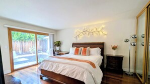 A bedroom with a large bed, a sliding glass door to a patio, a small table with a plant, and a mirror on the wall.
