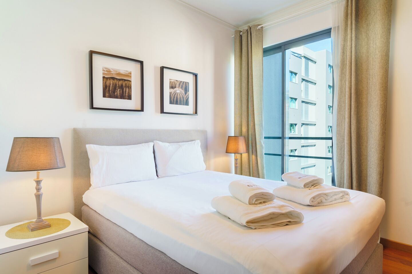 Habitación de hotel con una cama, dos cuadros en la pared, una mesita de noche con lámpara y una ventana con cortinas.